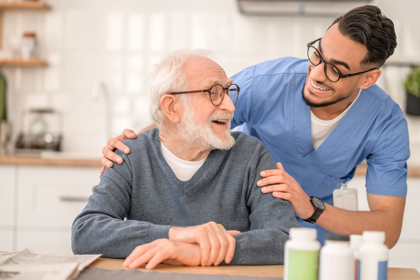 Smiling male nurse in uniform hugging a happy aged man seated at the kitchen table